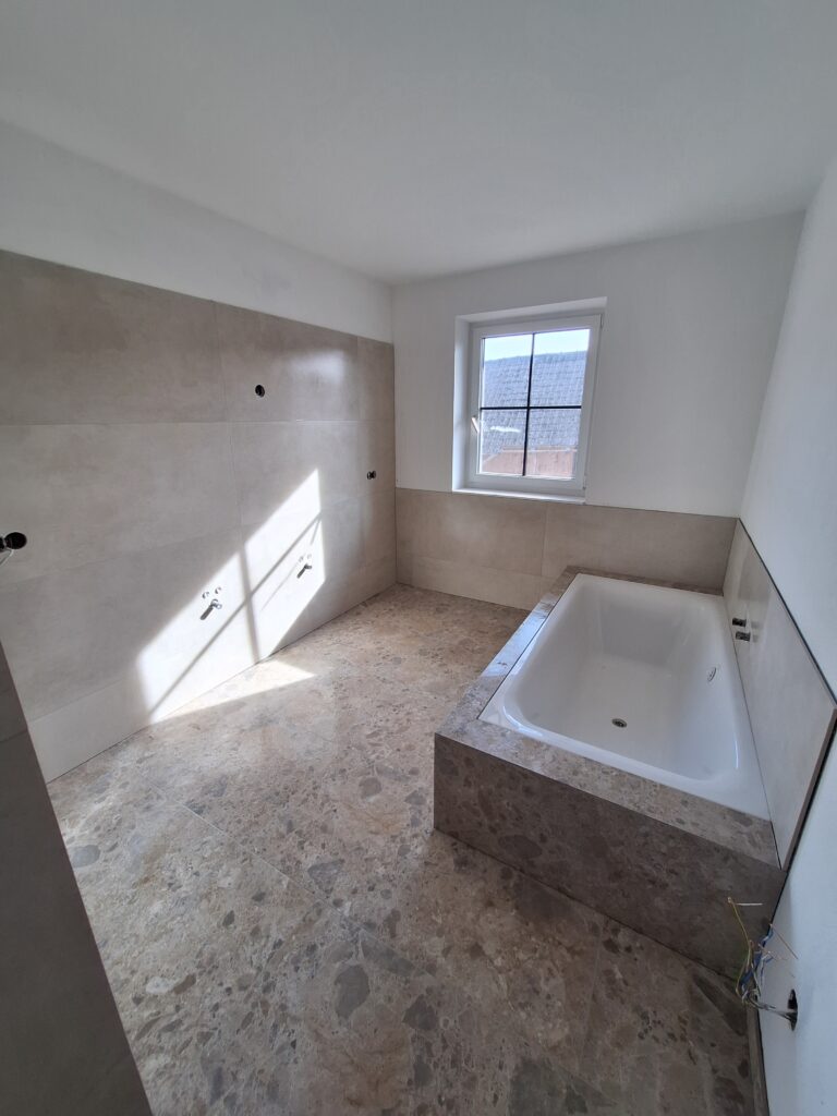Bad mit großformatigen Wandfliesen, Boden in Terrazzo-Optik und verfliester Wanne unter dem Fenster